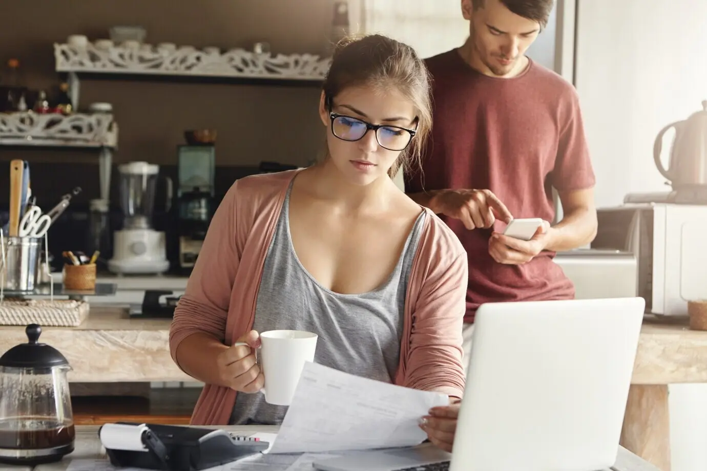 Красивая серьёзная молодая женщина в прямоугольных очках пьёт кофе и изучает счёт в руке, сидя в современном кухонном интерьере перед открытым ноутбуком.